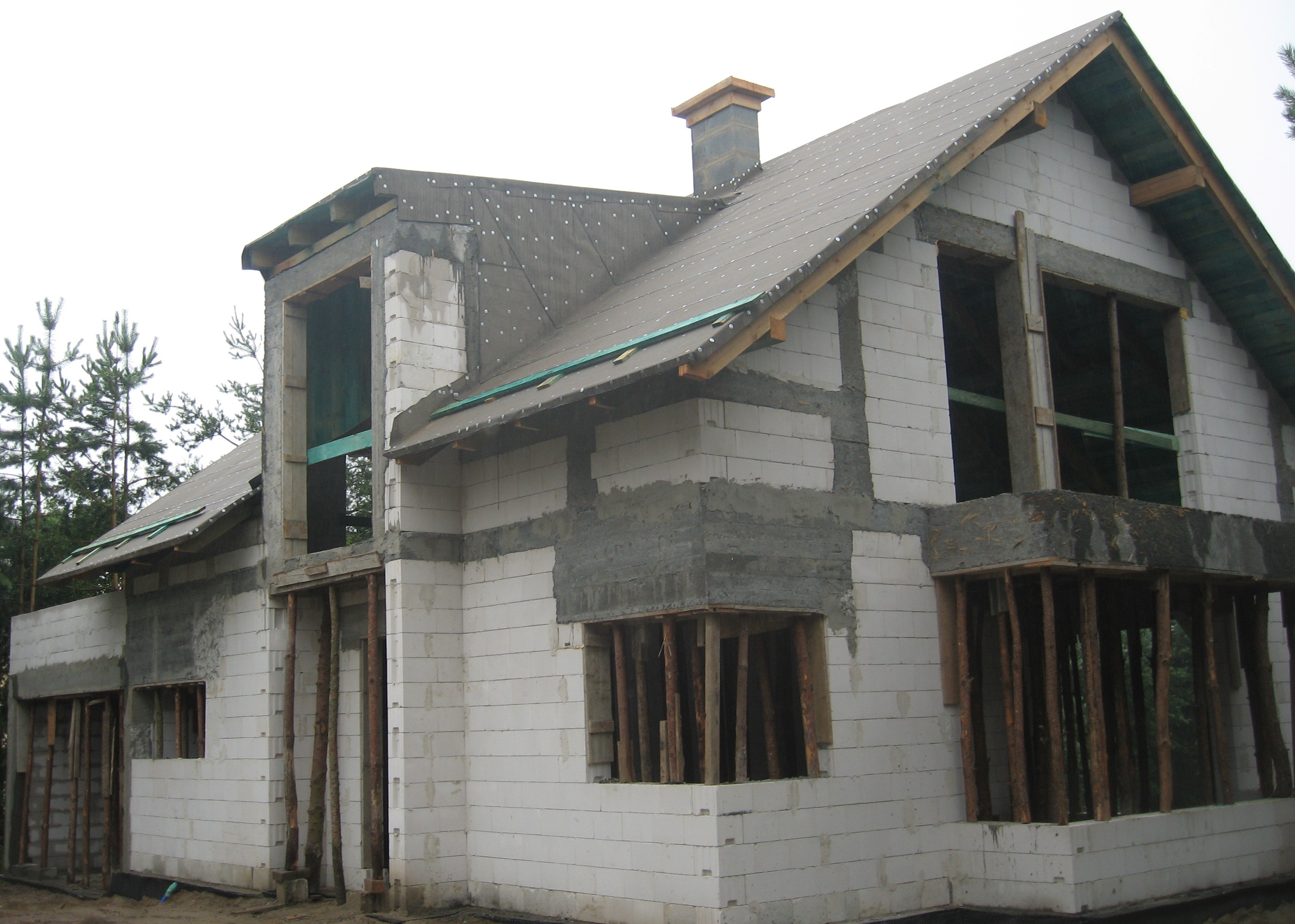 Surowy stan domu z białych bloczków, widoczne zbrojenia i szalunki okienne, dach pokryty papą, komin w trakcie budowy, w tle drzewa.