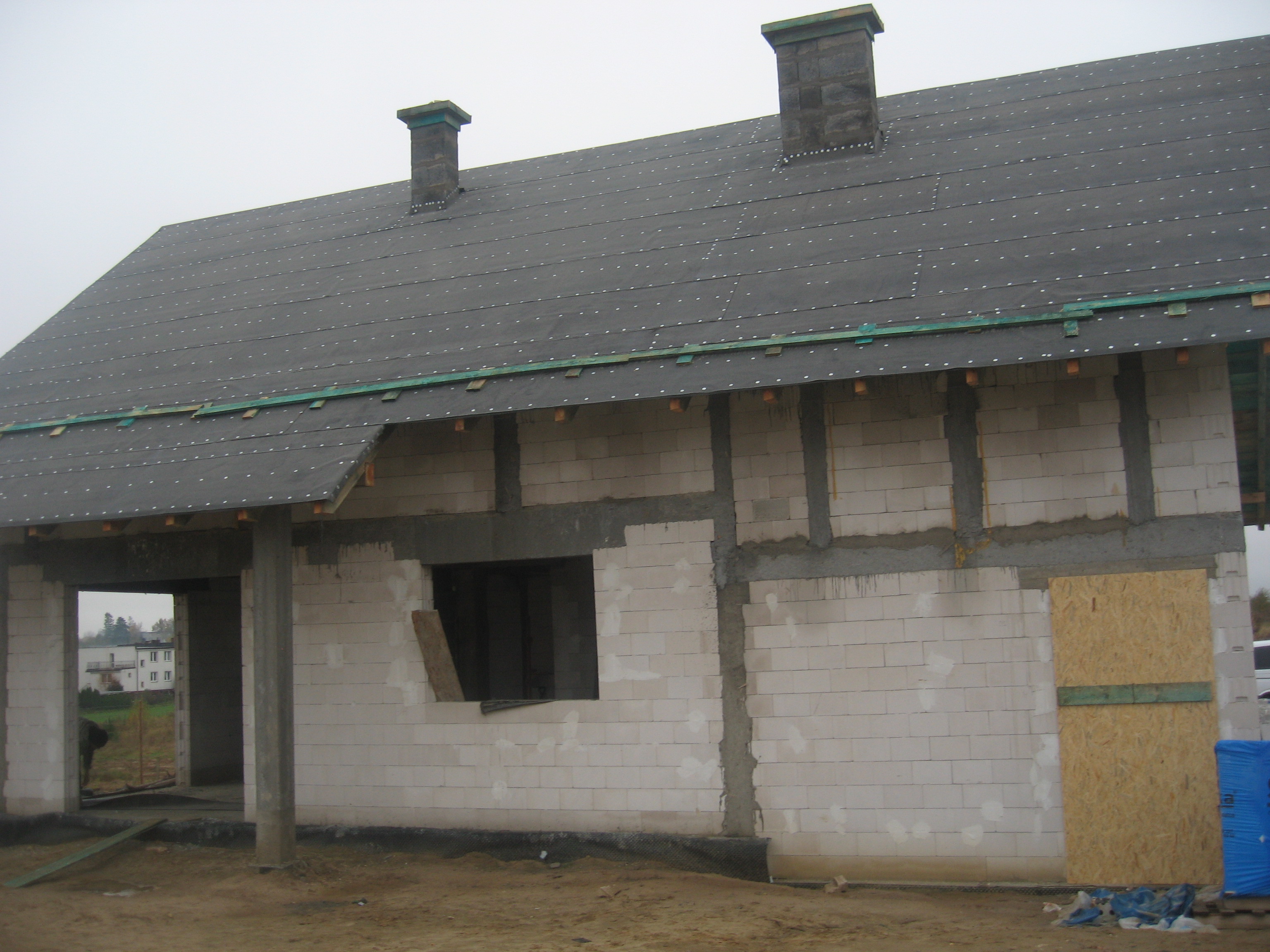 Surowy stan domu z białych bloczków, z częściowo pokrytym dachem papą i dwoma kominami, widoczne elementy konstrukcyjne i zabezpieczenia okien sklejką.