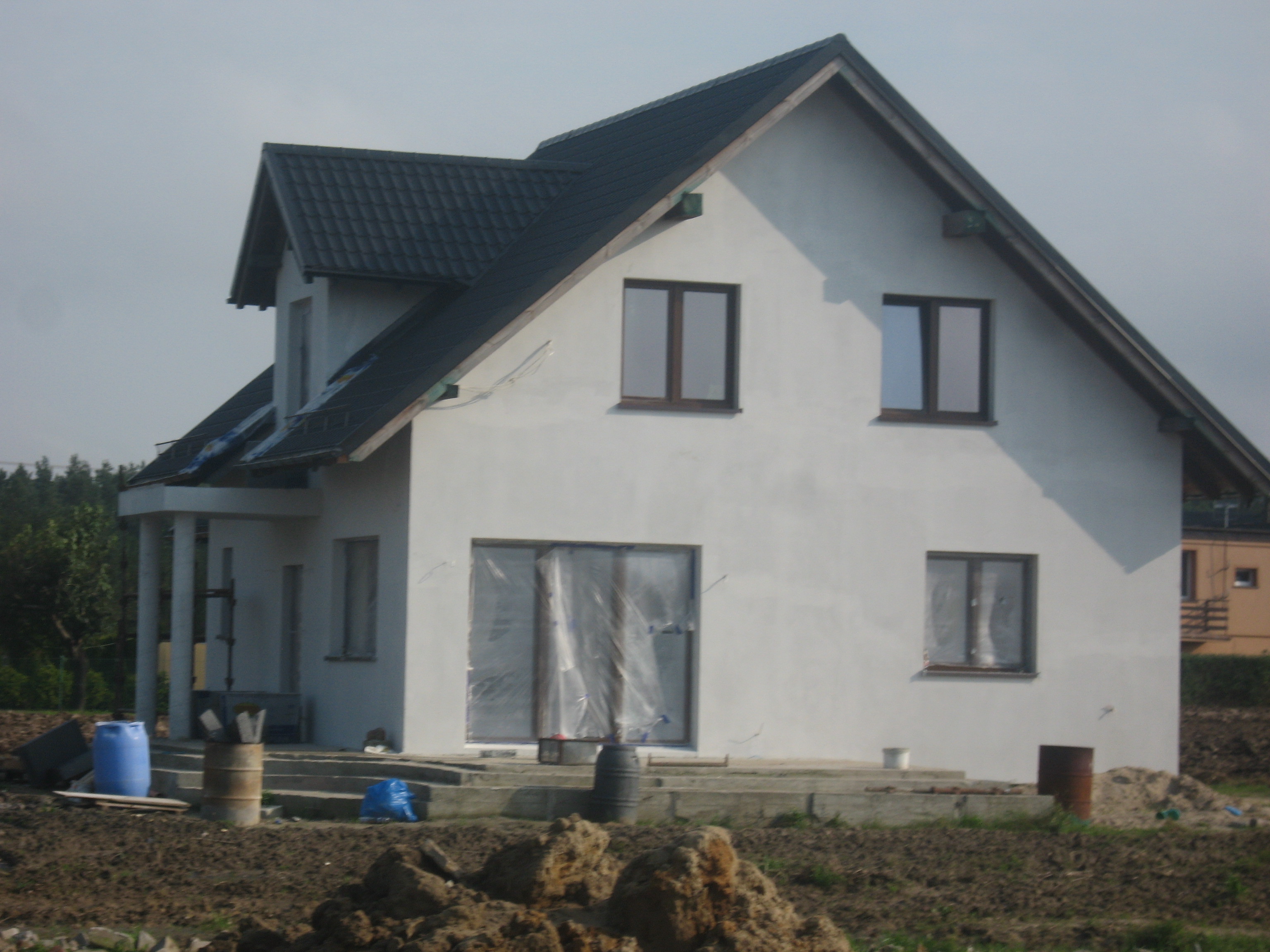 Surowy stan domu jednorodzinnego z ciemną dachówką, częściowo osłonięte folią okna, niezagospodarowany teren wokół.