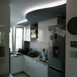 Nowoczesna kuchnia z białymi szafkami, szarą falowaną podsufitką z oświetleniem LED, srebrną lodówką z magnesami i zegarem na ścianie.