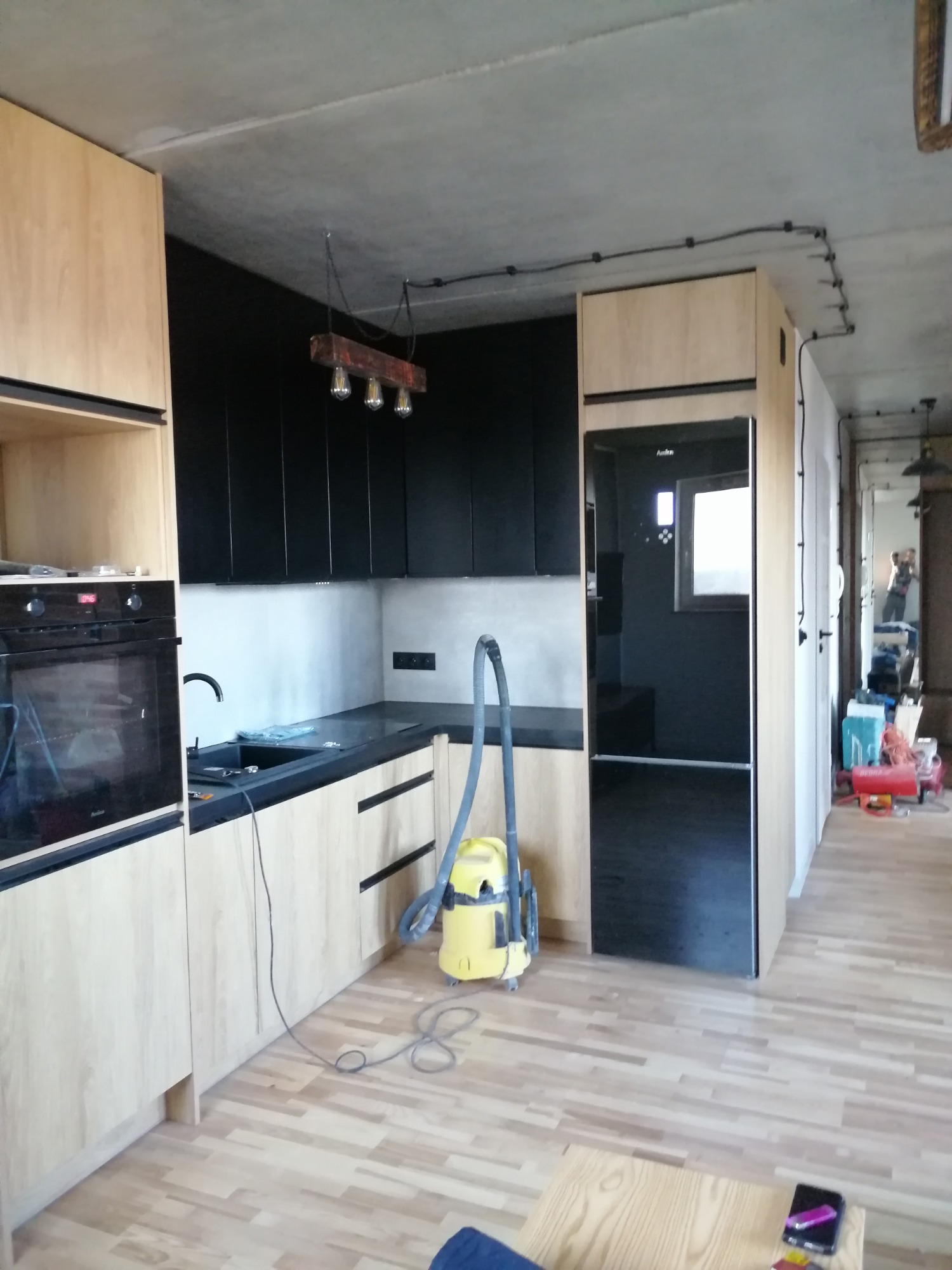 Nowoczesna kuchnia z drewnianymi szafkami i czarnymi frontami, surowy betonowy sufit z widocznymi instalacjami elektrycznymi, żółty odkurzacz na podłodze z paneli, widok na dalszą część mieszkania.