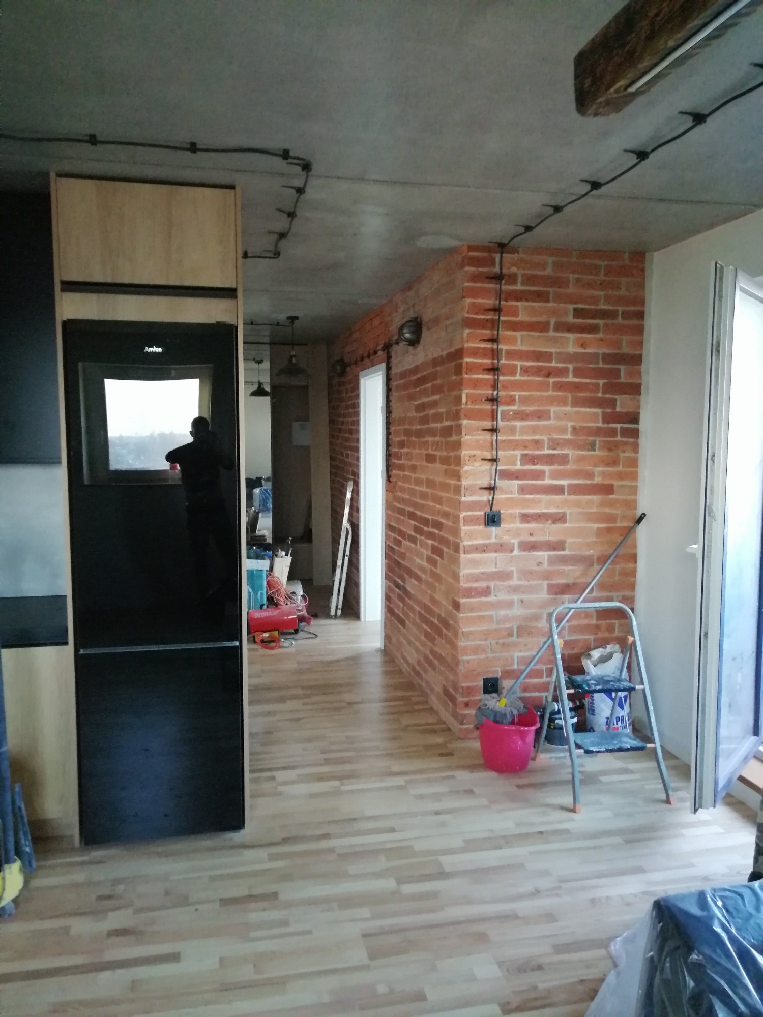 Wnętrze mieszkania w trakcie prac wykończeniowych: widoczna ściana z czerwonej cegły z poprowadzonymi kablami elektrycznymi po wierzchu, drewniana podłoga, drabina malarska, lodówka z czarnym...
