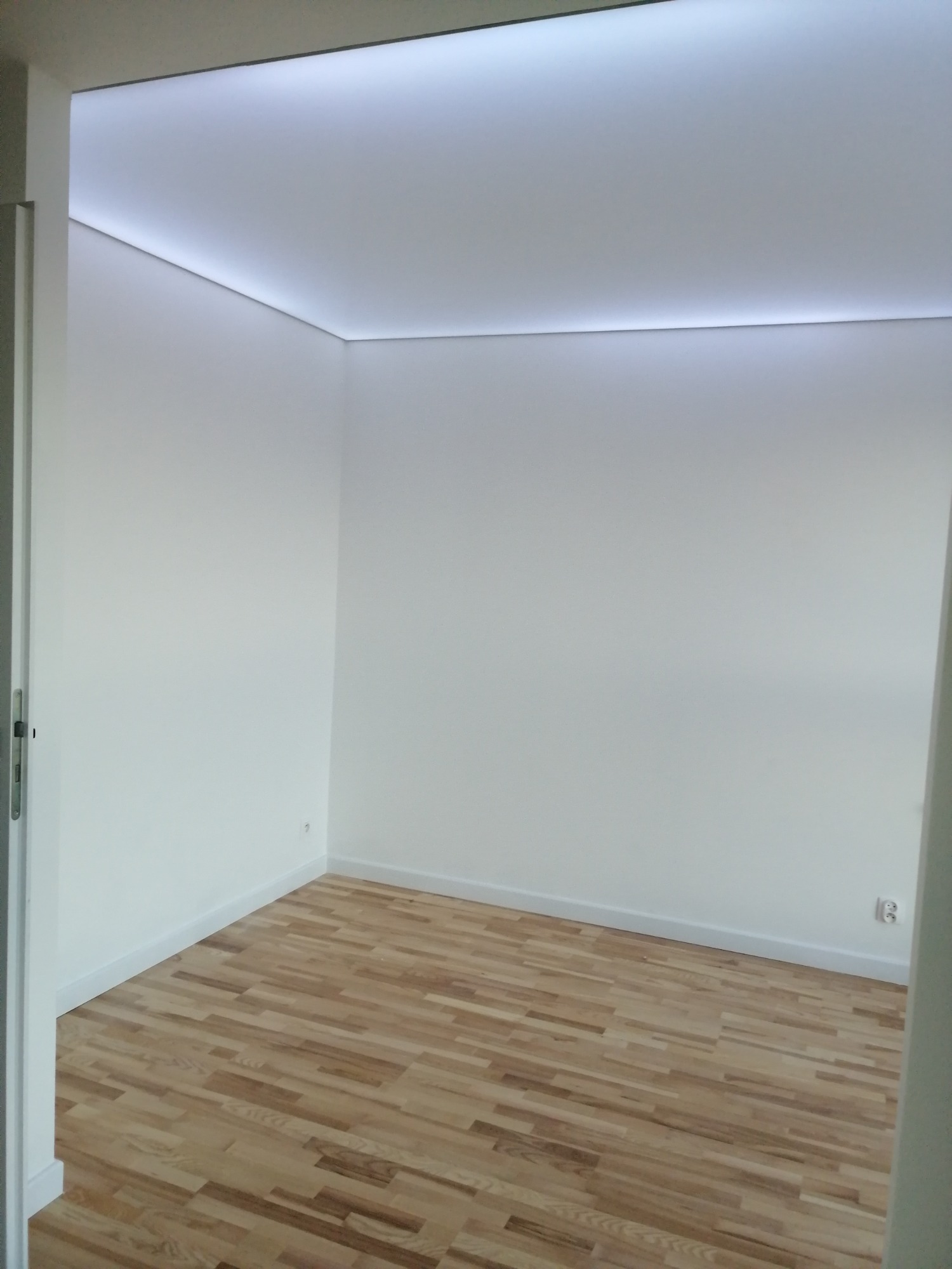 Pusty, odnowiony pokój z parkietem ułożonym w jodełkę i białymi ścianami z subtelnym oświetleniem LED wzdłuż sufitu.