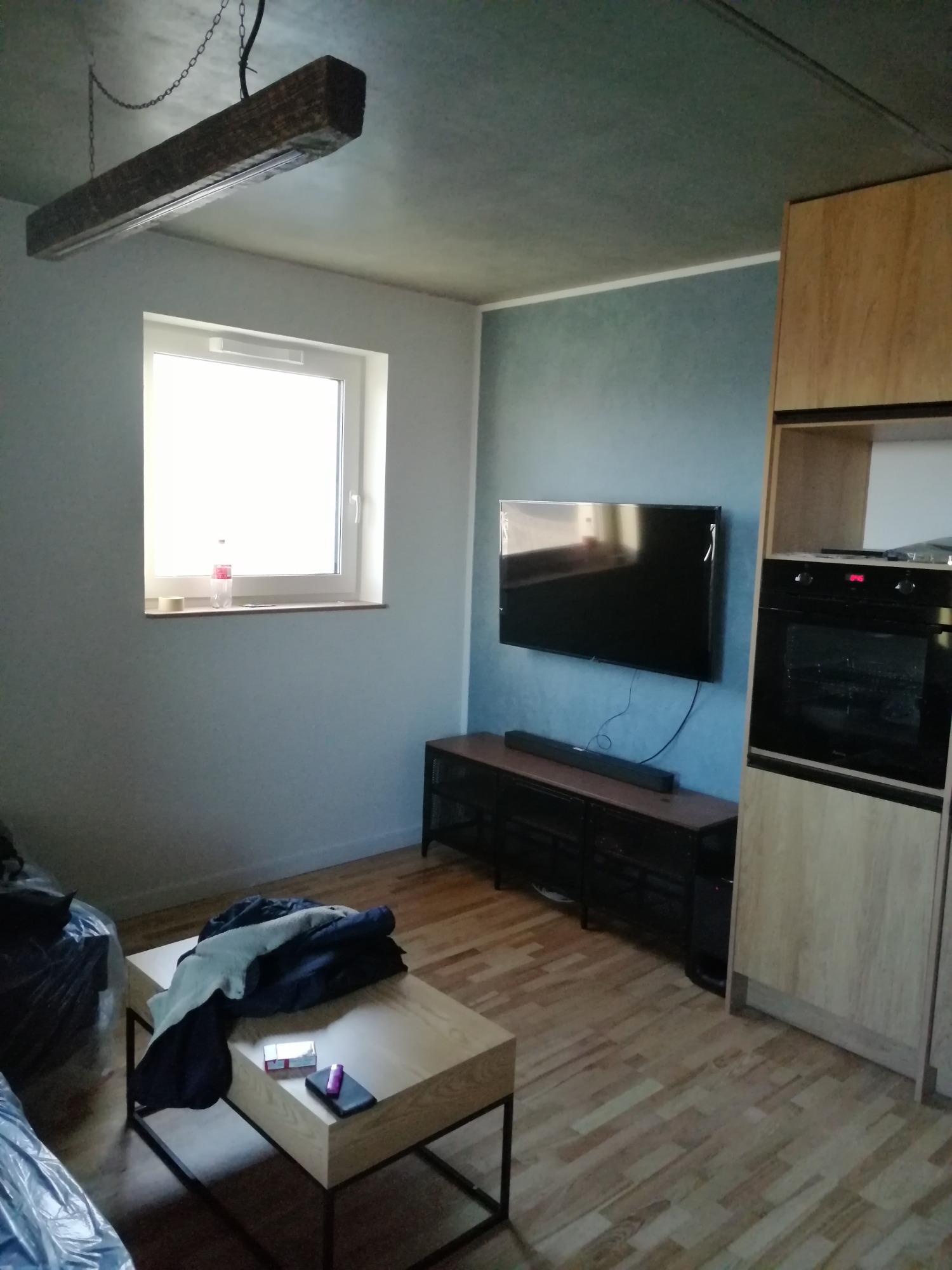 Wnętrze pokoju po remoncie: drewniana podłoga, niebieska ściana z wiszącym telewizorem, drewniane szafki kuchenne z piekarnikiem, stolik kawowy z ubraniami i przedmiotami, oraz lampa sufitowa...
