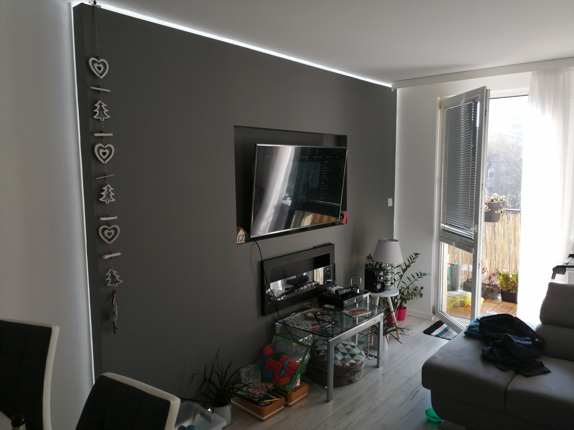 Szara ściana z podświetleniem LED, wnęką na telewizor i elektrycznym kominkiem, dekoracyjna girlanda z serduszkami i choinkami, balkon z roślinami w tle.