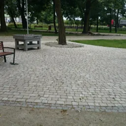 Wyłożona kostką brukową przestrzeń rekreacyjna z ławką, stołem do ping-ponga i grillem. Widoczne drzewa i zieleń w tle. Projekt zagospodarowania terenu.