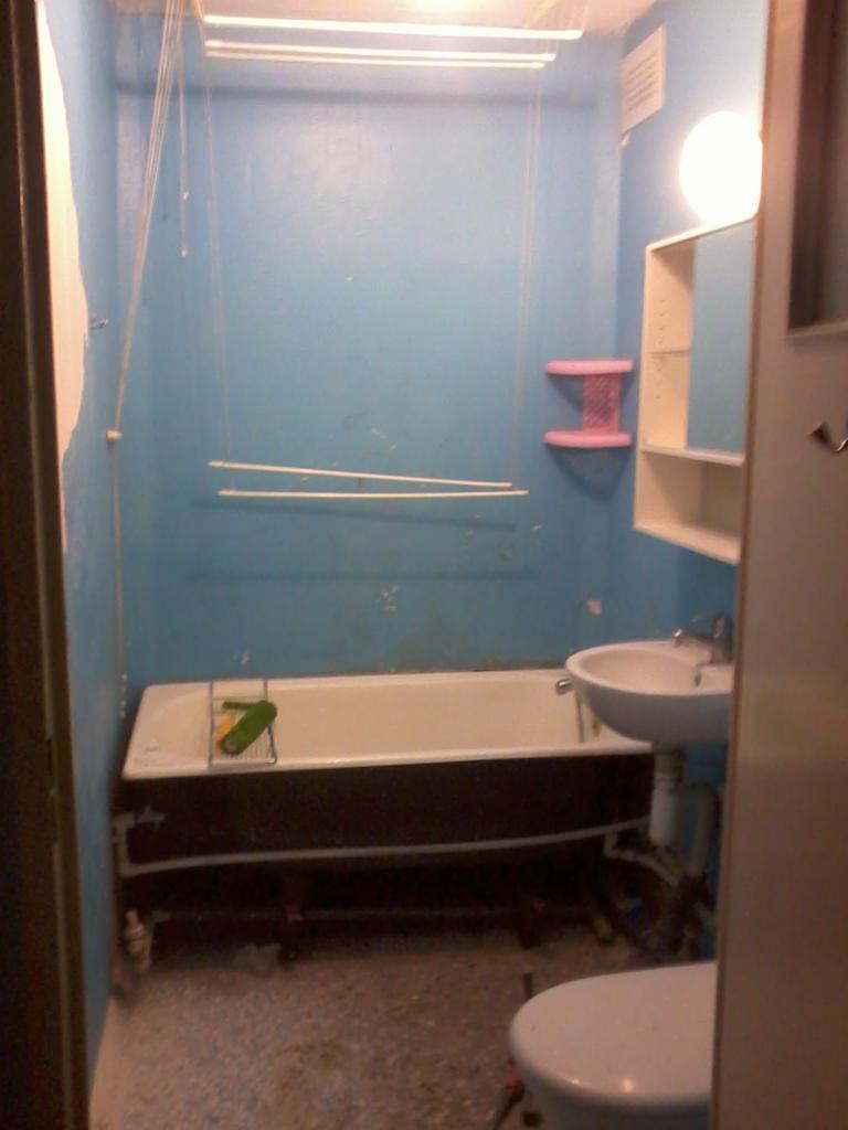 Niefachowo wykończona łazienka z niebieskimi ścianami, wanną i umywalką, widok z perspektywy drzwi. Na wannie leży gąbka.