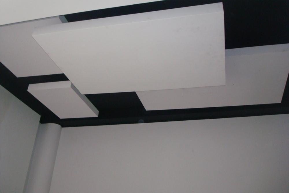 Sufit podwieszany z białych paneli o różnych kształtach i rozmiarach, zamontowanych na czarnej konstrukcji, widoczny fragment ściany pomalowanej na biało oraz kolumna.