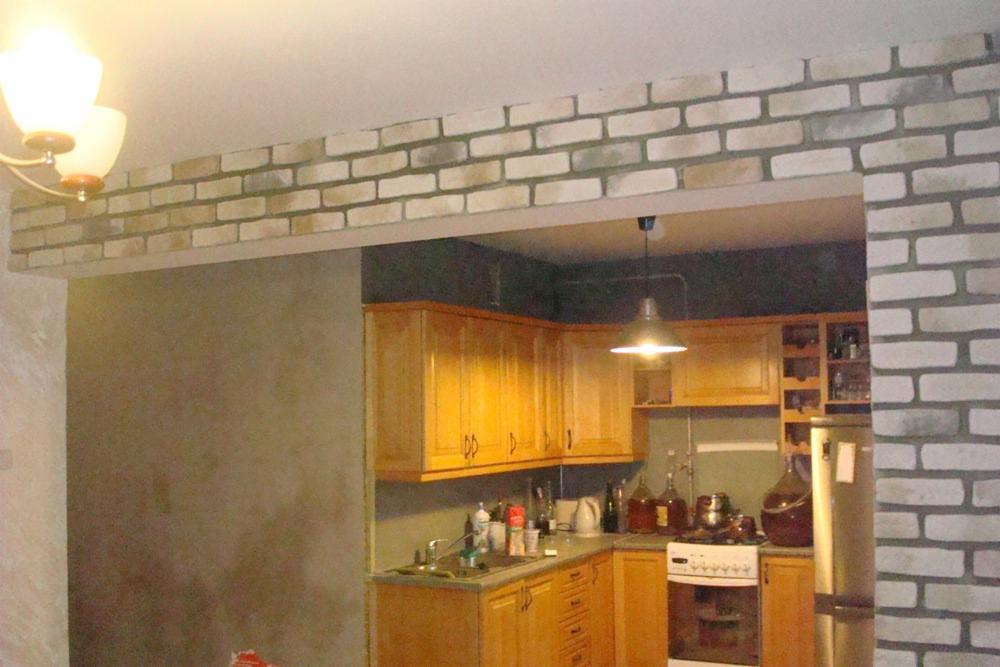 Wykończone przejście między pokojami z dekoracyjną cegłą w odcieniach szarości i beżu, widok na kuchnię z drewnianymi szafkami i oświetleniem halogenowym.