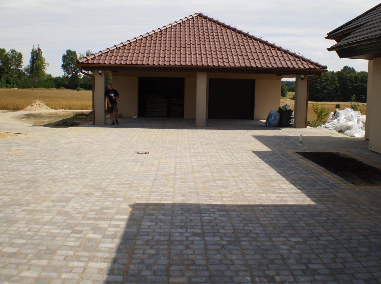 Jasna kostka brukowa ułożona na podjeździe przed garażem z czerwoną dachówką. Mężczyzna oparty o filar. Worki i hałda piasku w tle.