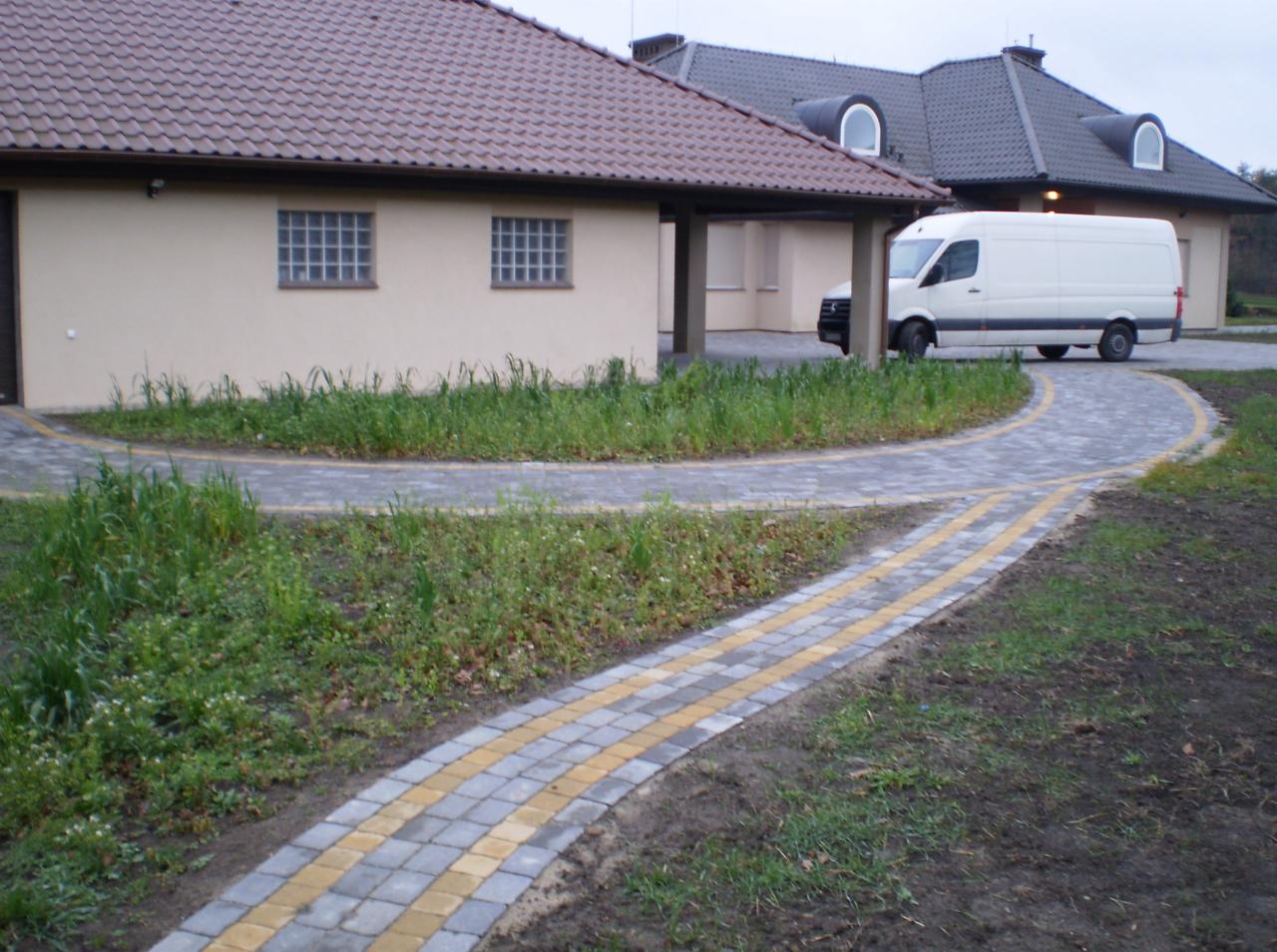 Ułożona z kostki brukowej ścieżka prowadzi do budynku z beżową elewacją i brązowym dachem. W tle biały van. Widoczne nierówności terenu i trawa.