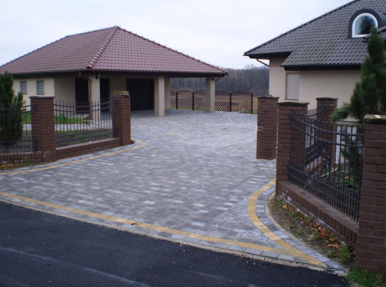 Ułożona kostka brukowa na podjeździe z widocznymi obrzeżami, ogrodzenie z ceglanymi słupkami i metalowymi przęsłami. Domy z brązowymi dachami w tle.