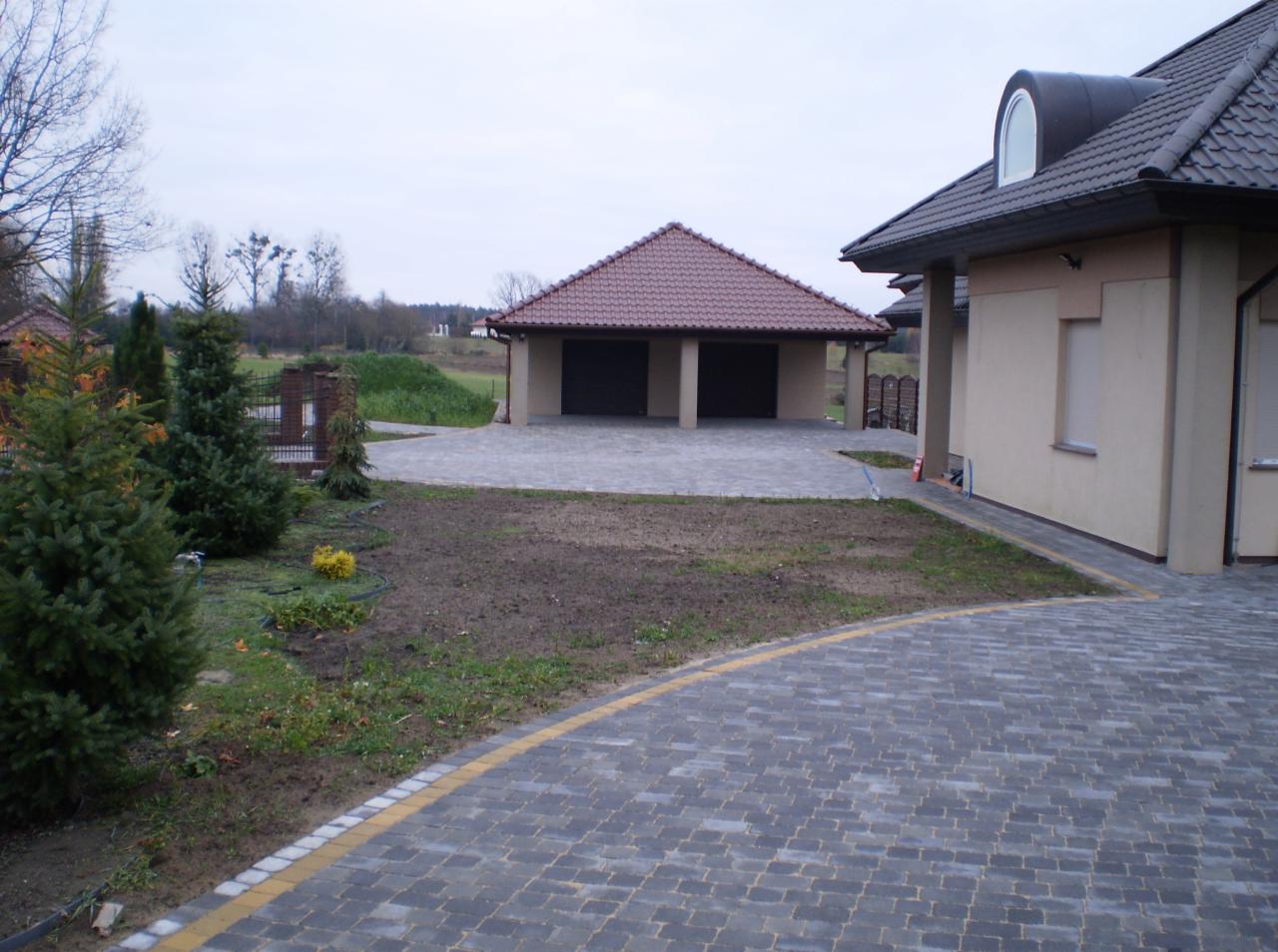Ułożona kostka brukowa w kolorze grafitowym z żółtą obwódką, prowadząca do garażu i domu z beżową elewacją. Widok z perspektywy ogrodu.