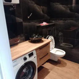 Nowoczesna łazienka z czarnymi płytkami imitującymi kamień, drewnianą szafką pod umywalkę, czarną umywalką nablatową, białą pralką Samsung i toaletą.