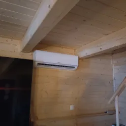 Biały klimatyzator Kaisai zamontowany na drewnianej ścianie w domu z boazerią, widoczny fragment sufitu i czarny telewizor. Oświetlenie naturalne.