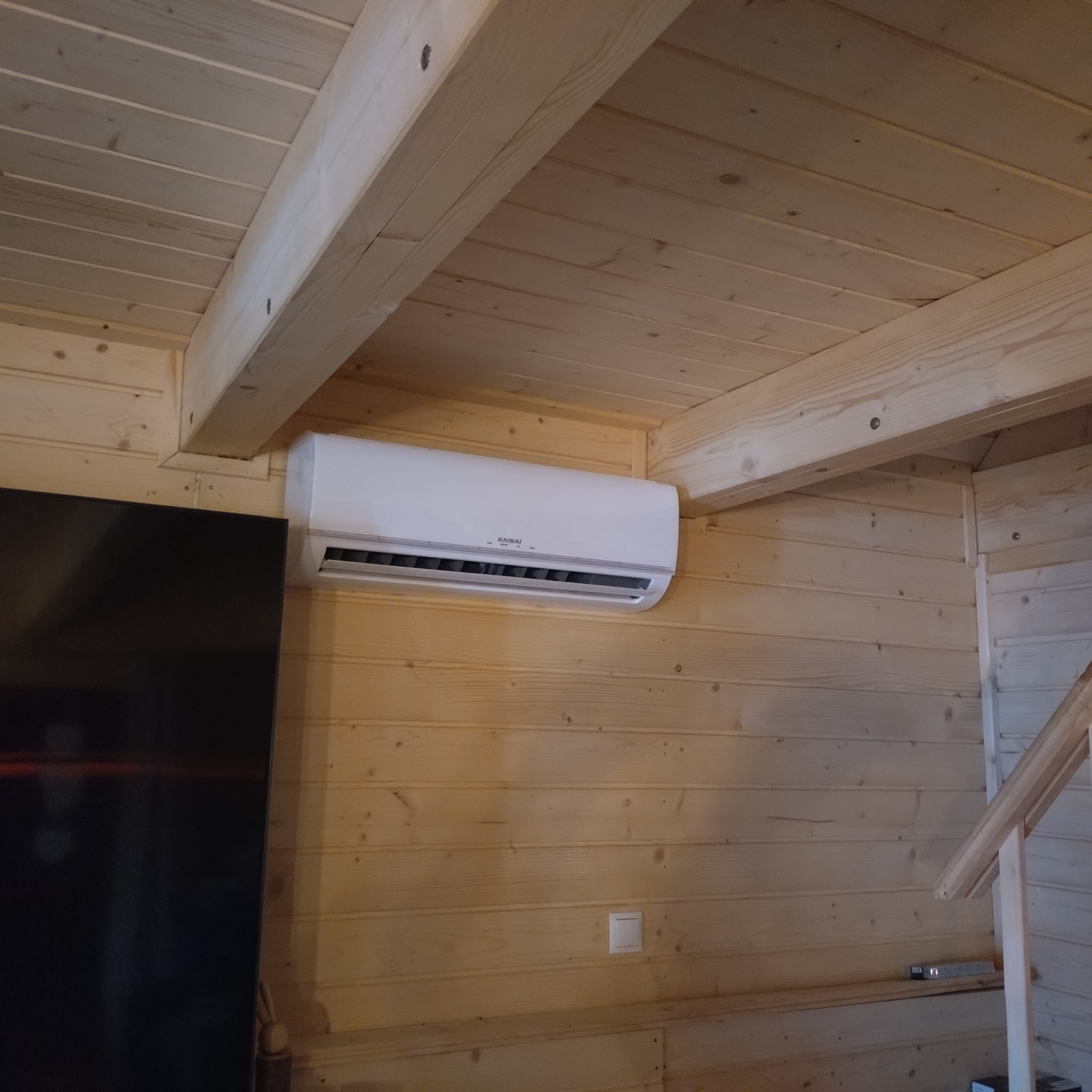 Biały klimatyzator Kaisai zamontowany na drewnianej ścianie w domu z boazerią, widoczny fragment sufitu i czarny telewizor. Oświetlenie naturalne.