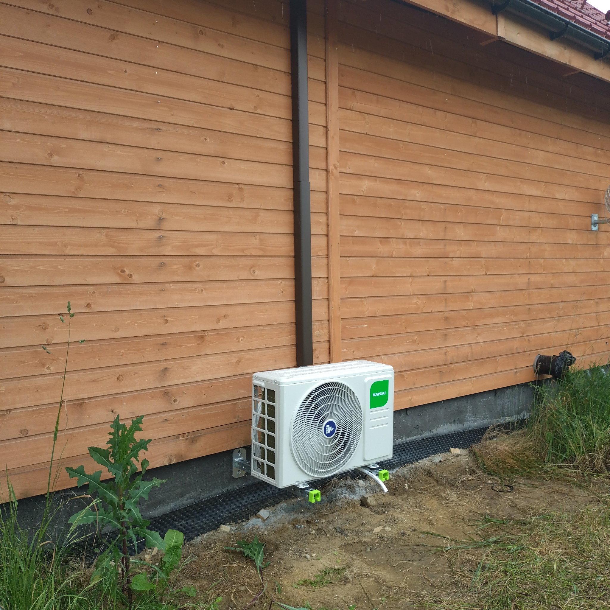 Biała jednostka zewnętrzna klimatyzacji Kaisai zamontowana na drewnianej elewacji domu, widoczne elementy montażowe i rurki. Trawa i ziemia w tle.