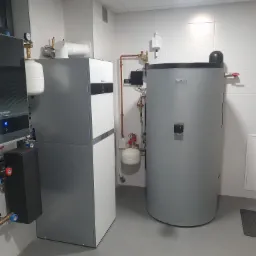 Kompaktowa kotłownia z szarą pompą ciepła Buderus, zbiornikiem akumulacyjnym i rozbudowaną instalacją hydrauliczną z zaworami VORTEX w pomieszczeniu wyłożonym białymi płytkami.