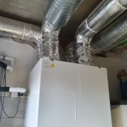 Biała centrala wentylacyjna z rekuperacją marki Vents, zamontowana pod sufitem z widocznymi rurami wentylacyjnymi w izolacji aluminiowej, na tle ściany z fragmentami starego tynku.