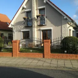 Dwukondygnacyjny dom z szarą elewacją, dekoracyjnymi balkonami z kwiatami, oraz ogrodzeniem z cegły i kutego żelaza w słoneczny dzień.