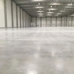 Wnętrze hali magazynowej o powierzchni 15000m2 w województwie mazowieckim z gładką, szarą posadzką, betonowymi kolumnami i szeregiem bram wjazdowych.