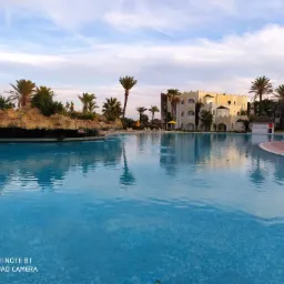 Rozległy basen w hotelu z dekoracyjną kaskadą wodną i palmami, w tle budynek hotelowy z elementami architektury mauretańskiej.