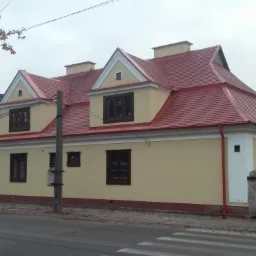 Odnowiony, parterowy budynek z czerwonym dachem, kremową elewacją i ciemnymi oknami. Widoczne detale architektoniczne i otoczenie miejskie.