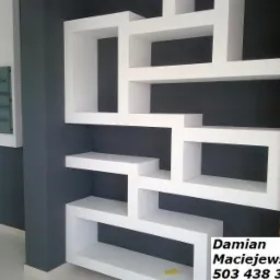 Biała, asymetryczna zabudowa z płyt gipsowych na ciemnej ścianie, z fragmentem ściany z cegły. Widoczny fragment podłogi z jasnych płytek.