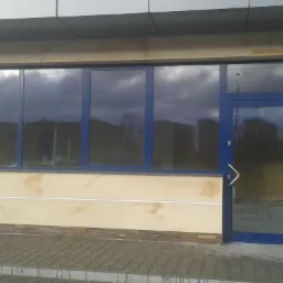 Elewacja budynku z oknami i drzwiami w niebieskich ramach. Dekoracyjna faktura ściany w odcieniach beżu i brązu. Widoczne elementy wykończeniowe.