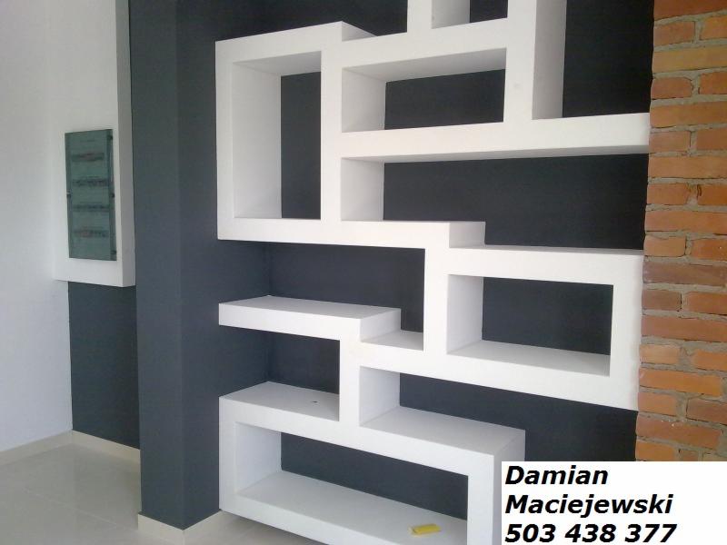 Biała, asymetryczna zabudowa z płyt gipsowych na ciemnej ścianie, z fragmentem ściany z cegły. Widoczny fragment podłogi z jasnych płytek.