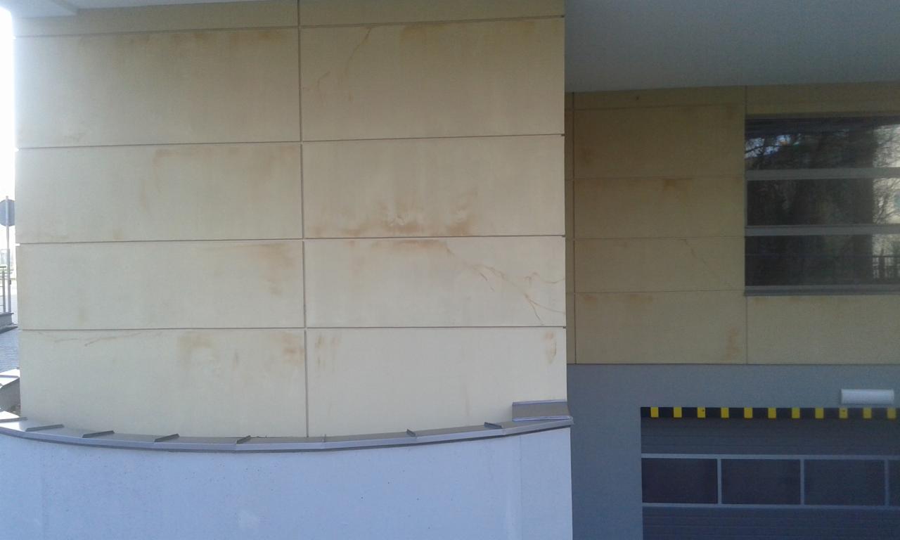 Fragment elewacji budynku z beżowymi płytami, widoczne zabrudzenia. Na drugim planie okno i brama garażowa z żółto-czarnymi pasami.