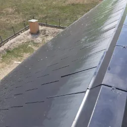 Nowe, czarne dachówki ułożone w rzędach, widoczne z góry pod kątem, z drewnianą budką w tle.