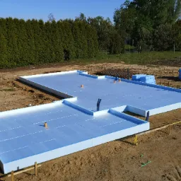 Płyta fundamentowa z izolacją XPS 200mm, widoczne pionowe rury instalacyjne i zbrojenie w tle, teren przygotowany pod budowę.