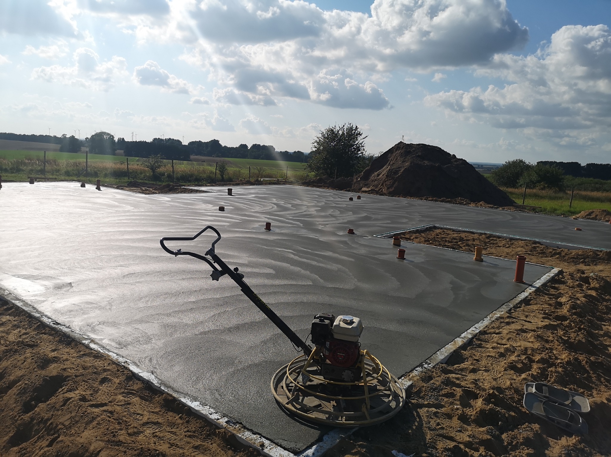 Świeżo wylana płyta fundamentowa, wygładzana zacieraczką mechaniczną, widoczne rurki instalacyjne, w tle usypana ziemia i krajobraz wiejski pod błękitnym niebem z chmurami.