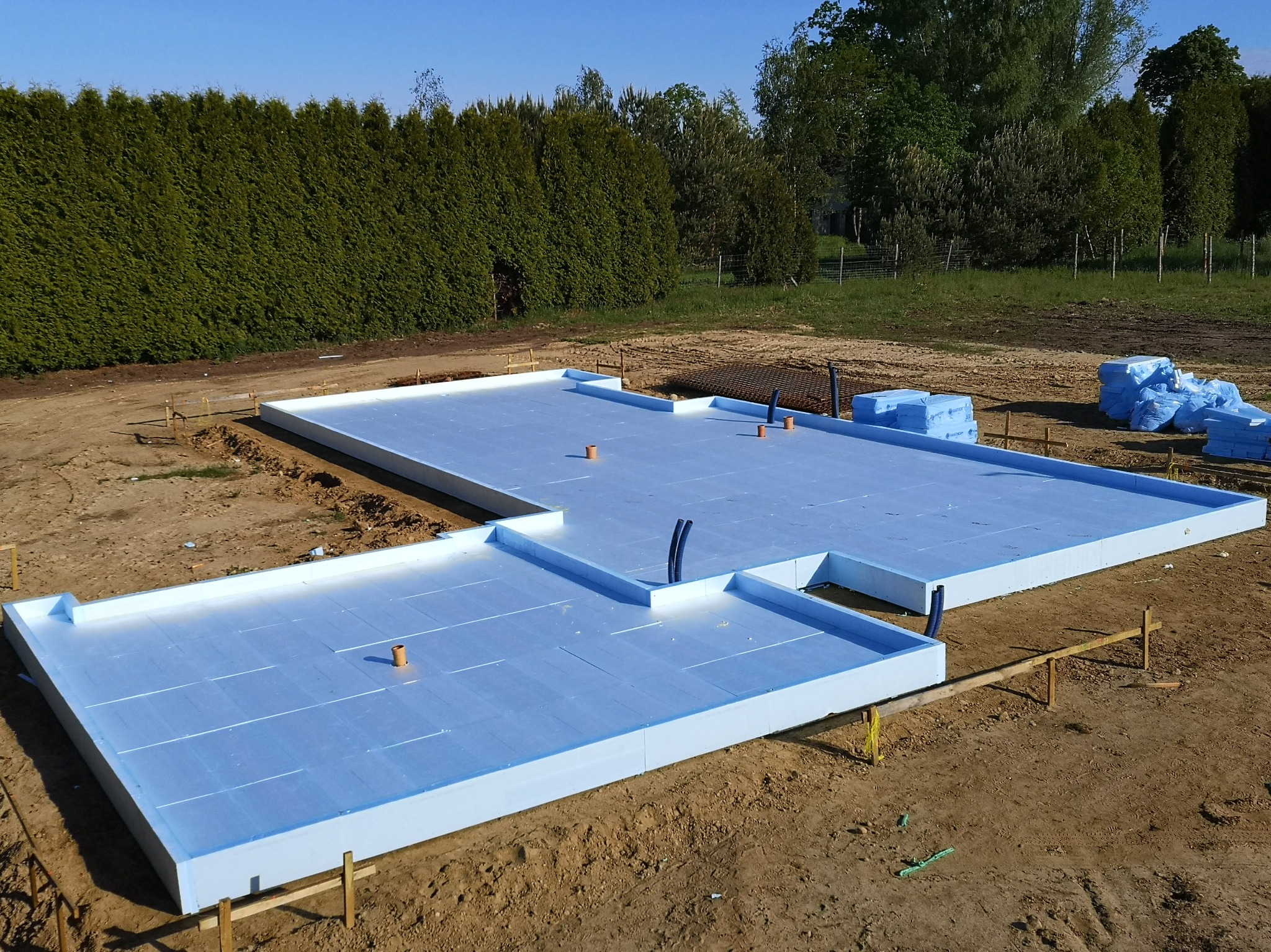 Płyta fundamentowa z izolacją XPS 200mm, widoczne pionowe rury instalacyjne i zbrojenie w tle, teren przygotowany pod budowę.