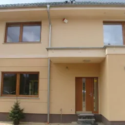 Dwukondygnacyjny dom z beżową elewacją, brązowymi oknami i drzwiami, oraz małą choinką przed jednym z okien. Widoczny fragment dachu i rynny.