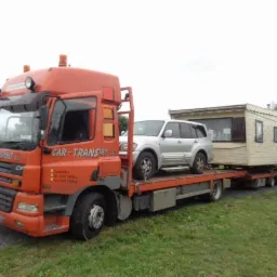 Pomarańczowa laweta z srebrnym SUV-em i przyczepą kempingową na platformie, zaparkowana na trawiastym terenie.