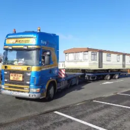 Niebiesko-złoty ciągnik siodłowy Scania z naczepą transportuje biało-beżowy domek mobilny na parkingu w słoneczny dzień.