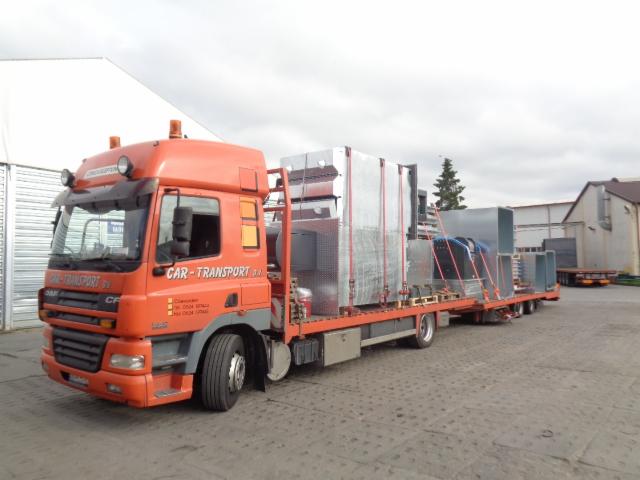 Pomarańczowa ciężarówka DAF z naczepą przewożąca elementy wentylacyjne zabezpieczone czerwonymi pasami, widok z boku na placu manewrowym.