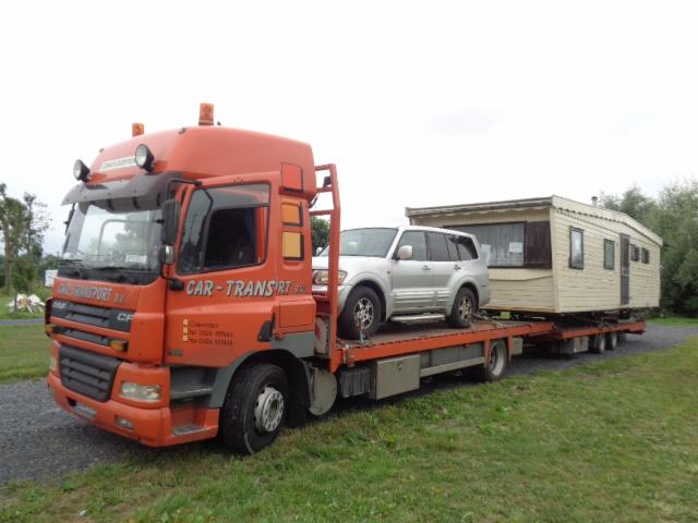 Pomarańczowa laweta z srebrnym SUV-em i przyczepą kempingową na platformie, zaparkowana na trawiastym terenie.