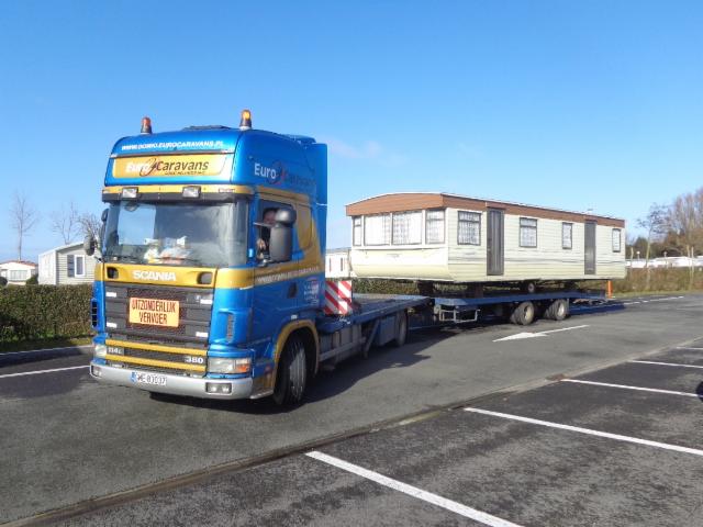 Niebiesko-złoty ciągnik siodłowy Scania z naczepą transportuje biało-beżowy domek mobilny na parkingu w słoneczny dzień.