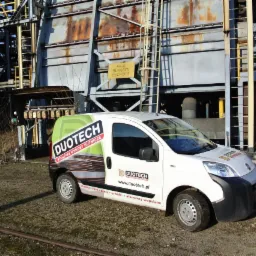 Biały samochód dostawczy firmy Duotech z logo i informacją o stalowych wyrobach hutniczych na tle konstrukcji przemysłowej z żółtą tabliczką informacyjną.