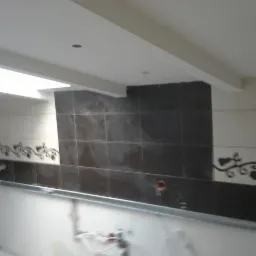 Fragment ściany w łazience z ułożonymi płytkami w odcieniach bieli i brązu, dekoracyjnym motywem roślinnym i widoczną instalacją. Wnętrze w trakcie wykończenia.