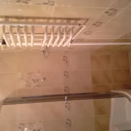 Biały grzejnik łazienkowy na ścianie z beżowych płytek, widoczny fragment kabiny prysznicowej. Ujęcie z dołu, podkreślające detale wykończenia.