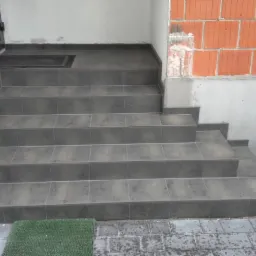 Wykończone płytkami schody wejściowe do budynku z ceglaną ścianą. Widoczne detale fug i wycieraczka. Płytki w odcieniach szarości.