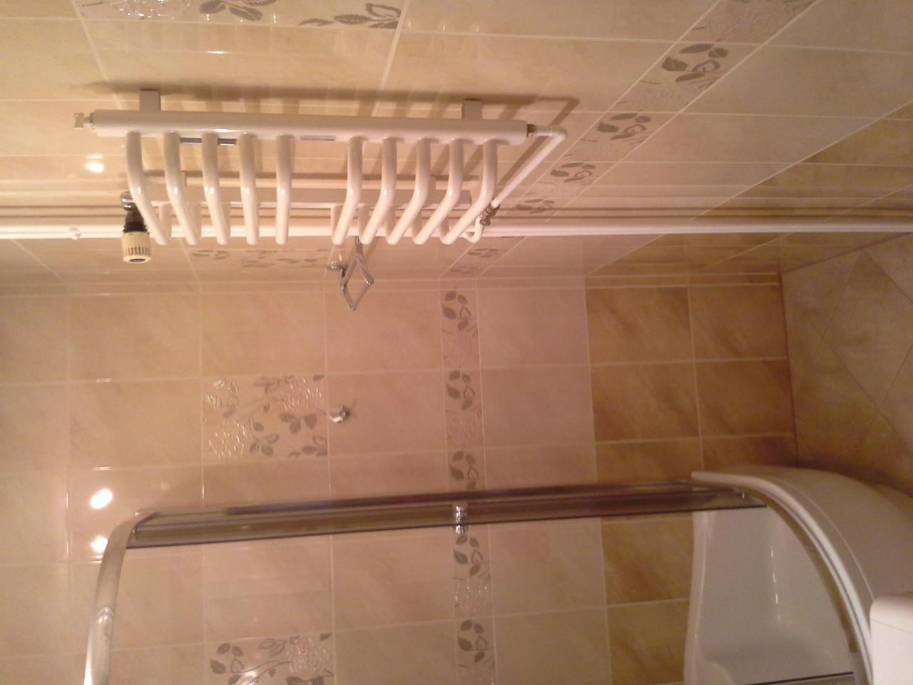 Biały grzejnik łazienkowy na ścianie z beżowych płytek, widoczny fragment kabiny prysznicowej. Ujęcie z dołu, podkreślające detale wykończenia.