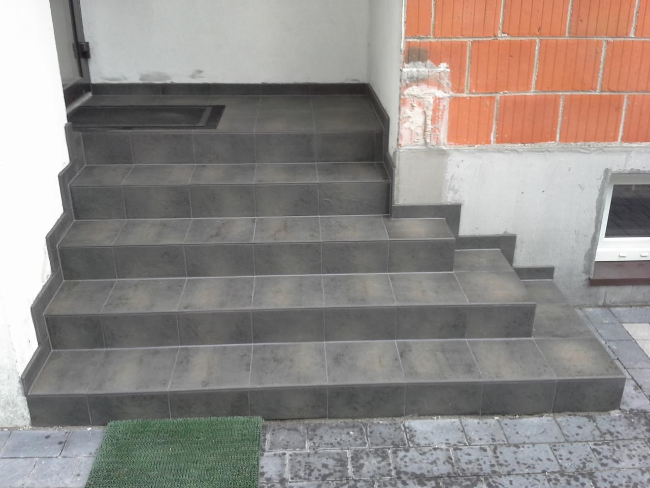 Wykończone płytkami schody wejściowe do budynku z ceglaną ścianą. Widoczne detale fug i wycieraczka. Płytki w odcieniach szarości.