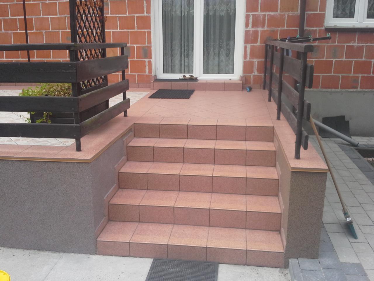 Wykończone schody wejściowe do domu z brązowych płytek, z drewnianą balustradą i wykończonym cokołem. Widoczne płytki na podeście i częściowo ułożona kostka brukowa obok.