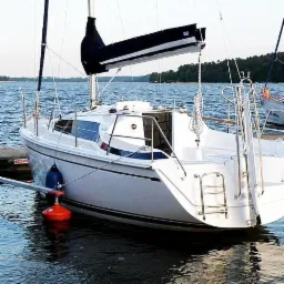 Biały jacht zacumowany przy pomoście, z widocznymi odbojnikami i drabinką rufową, na tle spokojnej wody i zielonego brzegu.