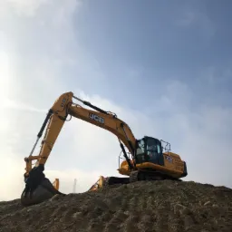 Żółta koparka JCB stoi na szczycie hałdy ziemi pod błękitnym niebem z delikatnymi chmurami, widoczny fragment drugiej maszyny w tle.